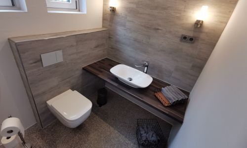 Modernes WC mit Hänge-WC, Aufsatzwaschbecken auf Holzplatte und stilvoller Wandbeleuchtung.